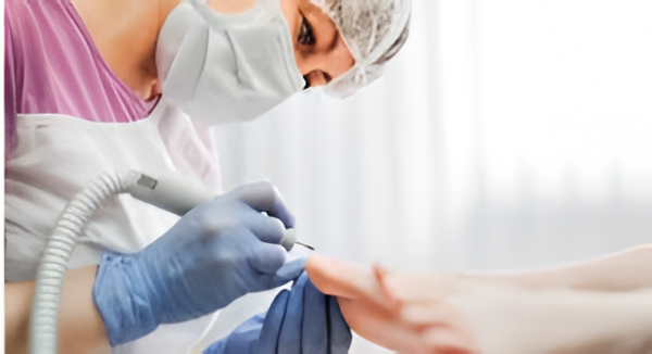 Profesional de la estética realizando un tratamiento de pedicura con un dispositivo eléctrico, usando guantes y mascarilla, en un ambiente iluminado y limpio.